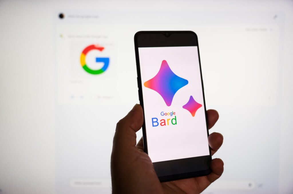 A hand holding a smartphone with the Bard logo is seen in front of a computer screen showing Google's logo. (Reuters Photo)