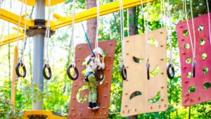 Gymnastic rings hang from ropes in a workout area as a young boy navigates a climbing panel in the background at an amusement park. (Shutterstock Photo)
