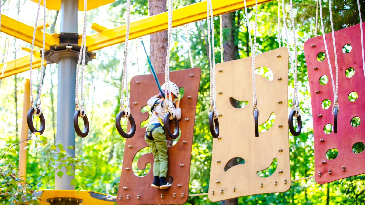 Gymnastic rings hang from ropes in a workout area as a young boy navigates a climbing panel in the background at an amusement park. (Shutterstock Photo)