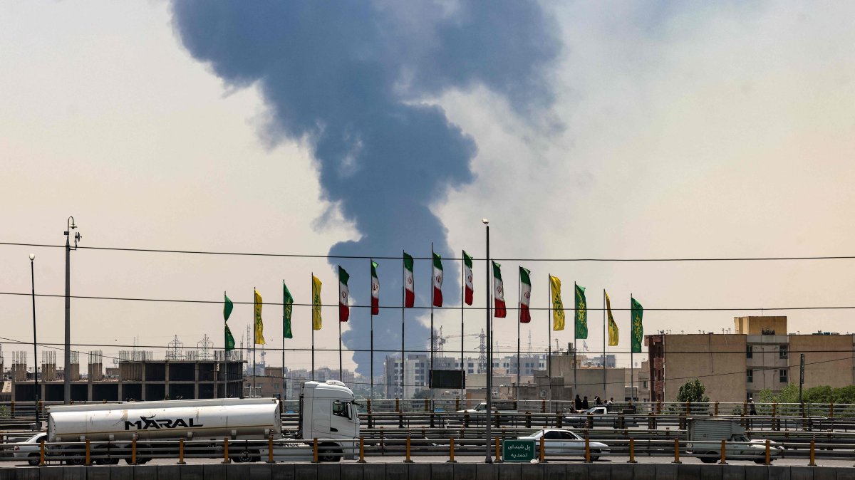 Flags flutter along a bridge as a plume of heavy smoke and fire rise from an oil refinery in southern Tehran, after it was hit in an overnight Israeli strike, June 15, 2025. (AFP Photo)